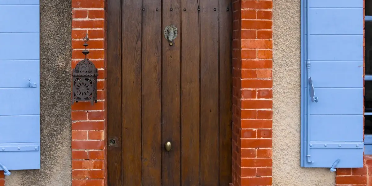 La façade d'une maison de village française : porte en bois, volets bleus et encadrements de brique sur un mur de pierre enduit.