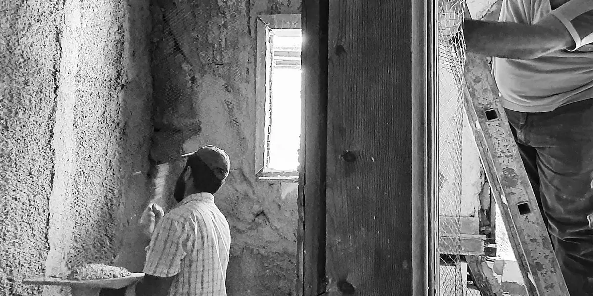 Deux artisans rénovent une pièce d'un bâtiment ancien : l'un applique un enduit sur un mur de pierre, l'autre pose un grillage depuis une échelle.